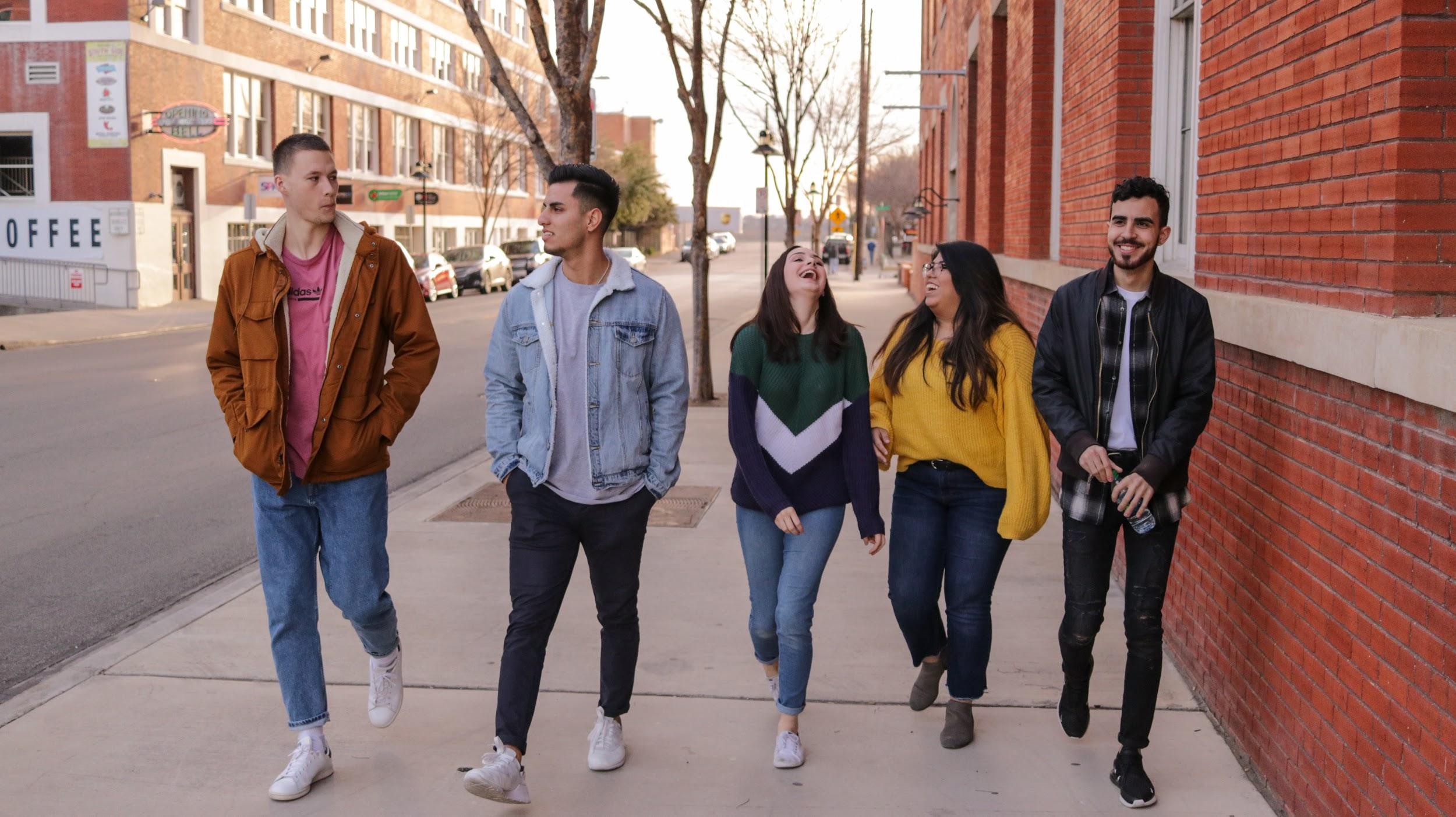 A group of teenagers (3 boys and 2 girls) walking down the city street laughing and talking.