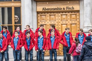 Two lines of 13 white male models, wearing the same outfit: jeans, a blue button down shirt, and a red sweater. They are standing in front of the doors of an Abercrombie and Fitch store.