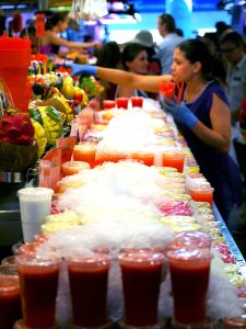 Market with colorful smoothies in plastic cups. A worker stands behind the smoothies handing them to customers with gloves on.